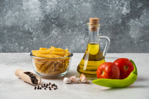 Pasta cruda en recipiente de vidrio, botella de aceite de oliva, granos de pimienta y verduras en el cuadro blanco.