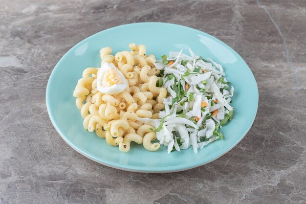 Pasta con ensalada fresca y huevo en placa azul.