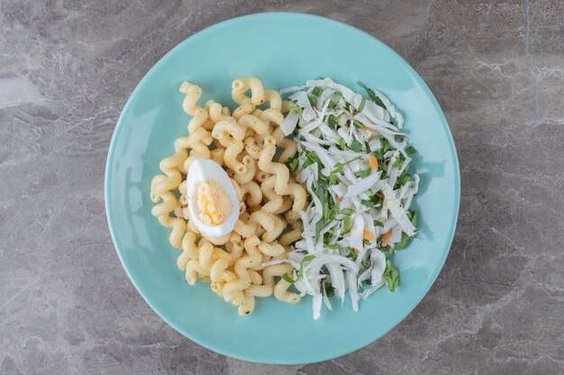 Pasta con ensalada fresca y huevo en placa azul.