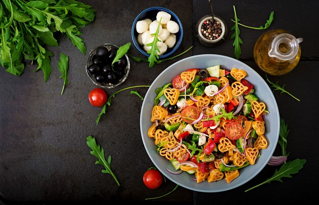 Pasta en forma de ensalada de corazón con tomates, pepinos, aceitunas, mozzarella y cebolla roja al estilo griego. Endecha plana. Vista superior