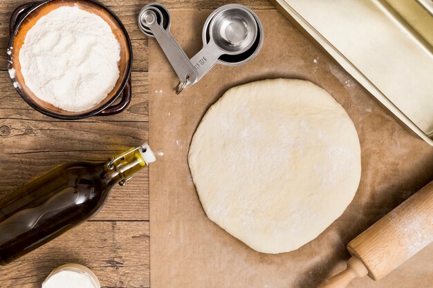 Pasta fresca lista para hornear en papel pergamino con cucharas de medir; harina; Aceite y rodillo sobre el escritorio de madera.