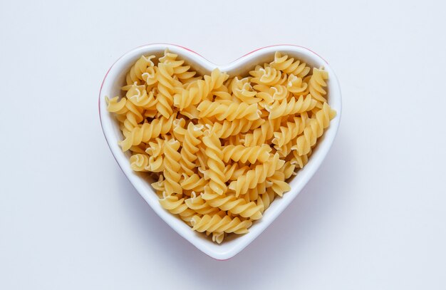 Pasta fusilli amarillo en un recipiente en forma de corazón sobre una mesa blanca. aplanada