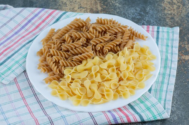 Pasta fusilli marrón y pastas farfalle en el cuenco sobre la toalla, sobre el fondo de mármol.