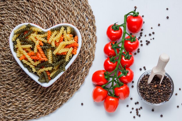 Pasta fusilli en un tazón con tomates, granos de pimienta en la vista superior primicia en blanco y mesa de mantel de mimbre