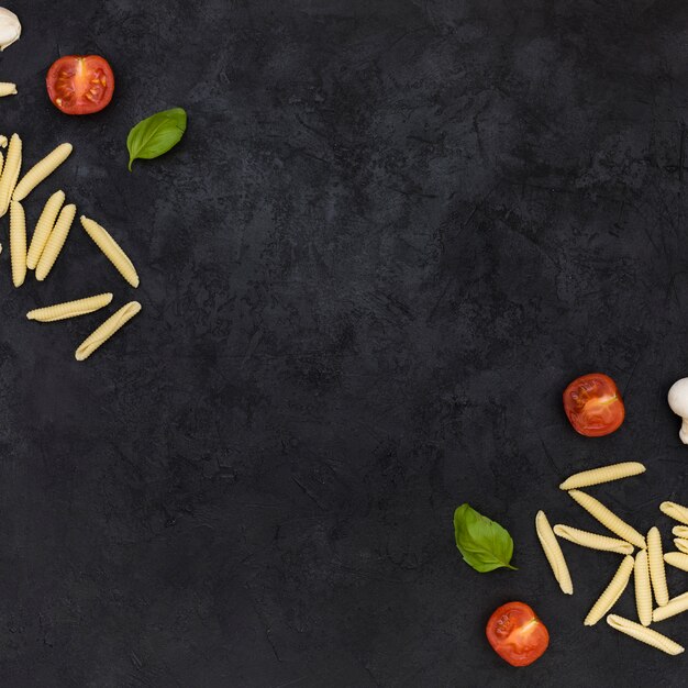 Pasta garganelli cruda con tomates a la mitad y albahaca en la esquina del fondo texturado negro