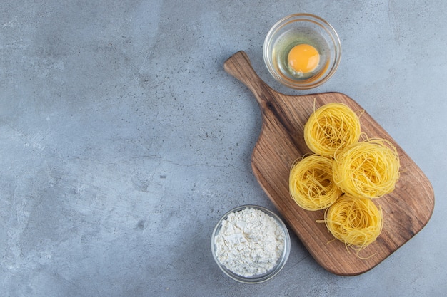 Pasta nido seca cruda con huevo crudo y un tazón de vidrio de harina sobre un fondo de piedra.