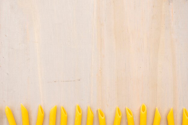 Pasta penne cruda amarilla dispuesta en fila sobre fondo con textura de madera