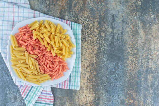 Pasta penne y fusilli en un recipiente sobre una toalla, sobre la superficie de mármol.