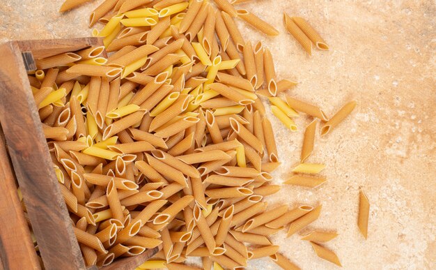 Pasta penne de grano entero en una caja de madera volcada, sobre el mármol.