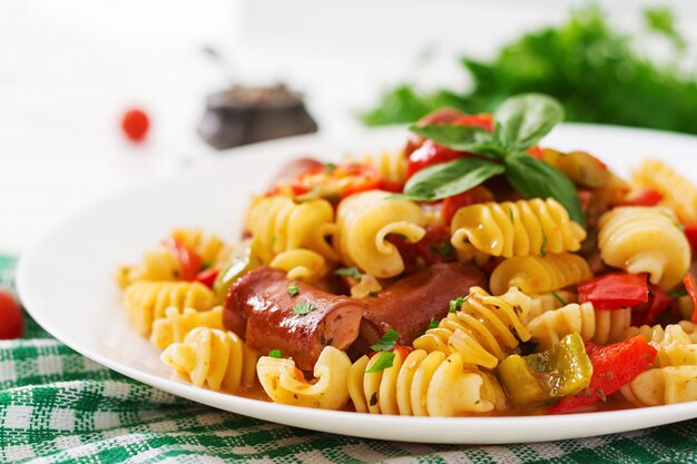 Pasta con salsa de tomate con salchicha, tomate, albahaca verde decorada en plato blanco sobre una mesa de madera.