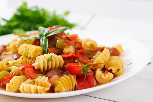 Pasta con salsa de tomate con salchicha, tomate, albahaca verde decorada en plato blanco sobre una mesa de madera.