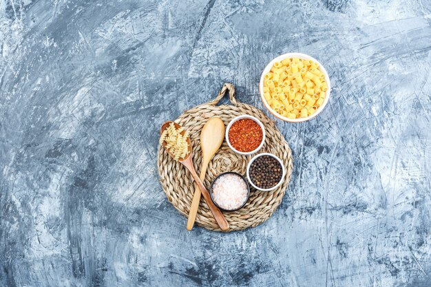 Pasta variada en un tazón y una cuchara de madera con especias vista superior sobre fondo gris de yeso y mantel de mimbre