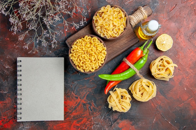 Pastas crudas, pimientos de cayena atados entre sí con una botella de aceite de cuerda, limón, ajo y cuaderno en la tabla de colores mezclados