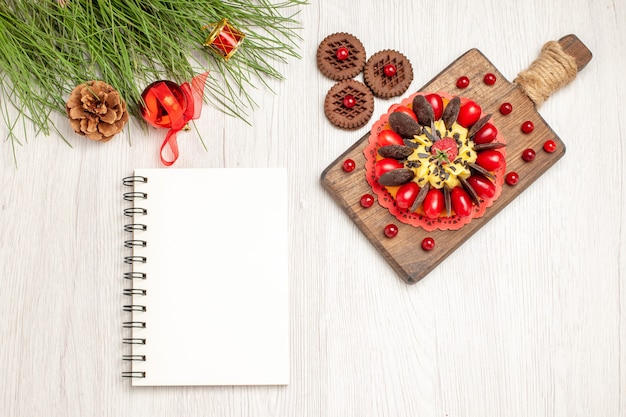 Pastel de bayas de vista superior en la tabla de cortar galletas y hojas de pino con juguetes navideños y un cuaderno en el suelo de madera blanca