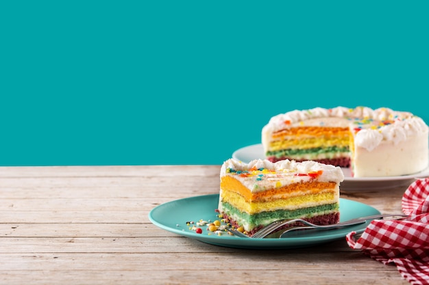 Pastel de capas de arco iris sobre mesa de madera y fondo azul.