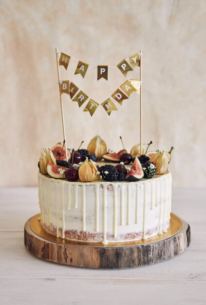 Un pastel de cumpleaños afrutado con adorno de cumpleaños, frutas en la parte superior y goteo blanco sobre fondo beige