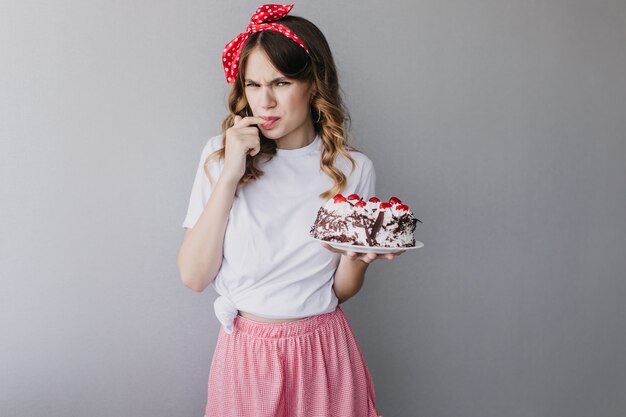 Pastel de degustación de mujer rizada divertida. Foto de adorable chica europea con lazo rojo en el pelo.