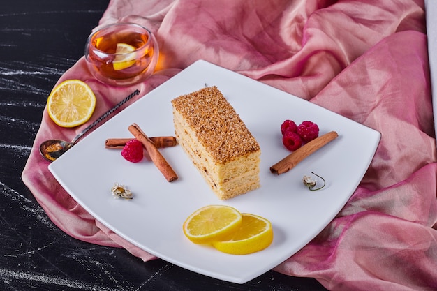 Pastel de miel con canela y frutas en plato blanco junto a mantel rosa.