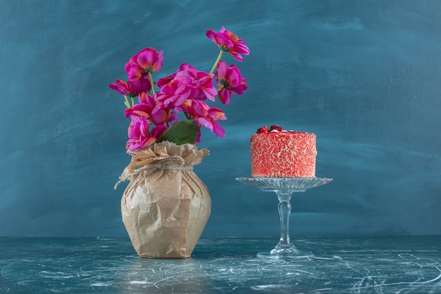 Pastel sobre pedestal junto a un jarrón de flores en azul.