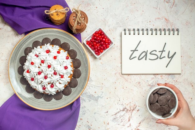 Pastel de vista superior con crema pastelera en la placa galletas de mantón morado atadas con bayas de cuerda en un tazón de fuente de chocolate en la mano femenina sabroso escrito en el bloc de notas en la mesa blanca