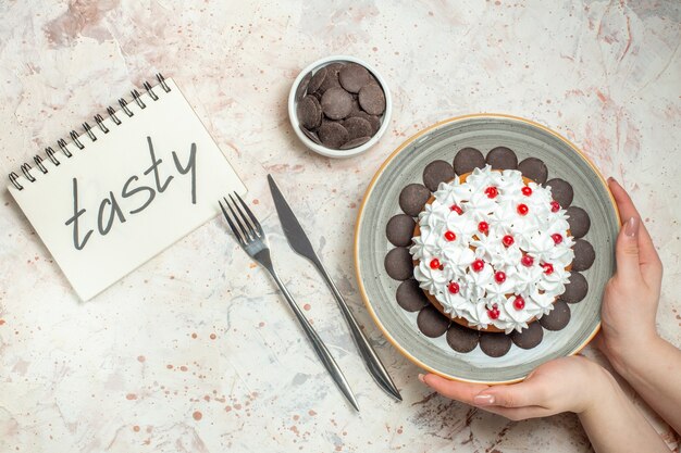 Pastel de vista superior con crema pastelera en placa ovalada en mano femenina chocolate en un tazón tenedor y cuchillo de cena sabroso escrito en el cuaderno