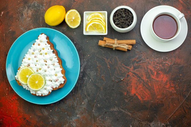 Pastel de vista superior con limón en placa azul rodajas de limón en un tazón taza de té palitos de canela sobre fondo rojo oscuro