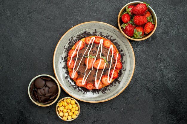 Pastel de vista superior con tazones de chocolate de fresa, avellana y chocolate alrededor del plato de pastel con chocolate y fresa en la mesa oscura