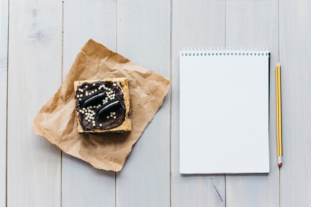 Pastelería fresca cerca de cuaderno de espiral y lápiz sobre tabla de madera