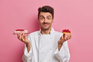 Foto gratis pastelero profesional trabaja en pastelería, sostiene deliciosos pasteles hechos a mano, posa en la cocina del restaurante, viste uniforme blanco