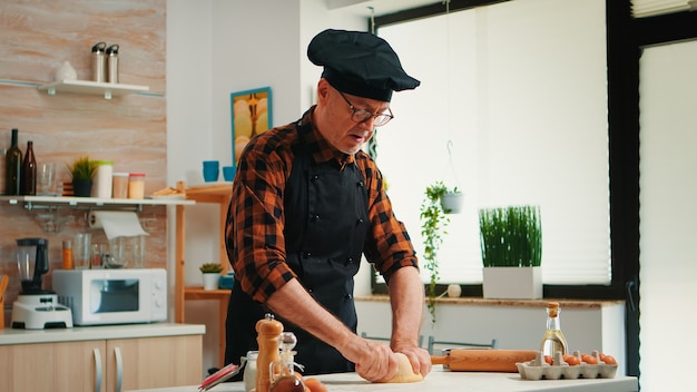 Foto gratuita pastelero trabajando en casa con masa cruda en la cocina moderna grabando la receta. anciano panadero jubilado con bonete mezclando ingredientes con harina tamizada amasando para hornear pan tradicional.