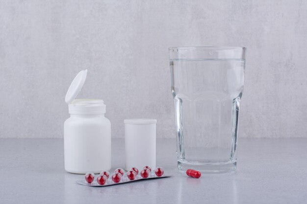 Pastillas rojas, recipientes y vaso de agua sobre la superficie de mármol. Foto de alta calidad