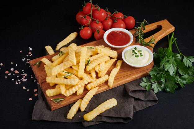 Patatas fritas caseras al horno con mayonesa, salsa de tomate y romero sobre tabla de madera. Sabrosas papas fritas en la tabla de cortar, en una bolsa de papel marrón sobre fondo de mesa de piedra negra, comida poco saludable.