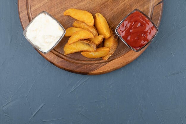 Patatas fritas con tazones de ketchup y mayonesa sobre tabla de madera en fondo azul. Foto de alta calidad