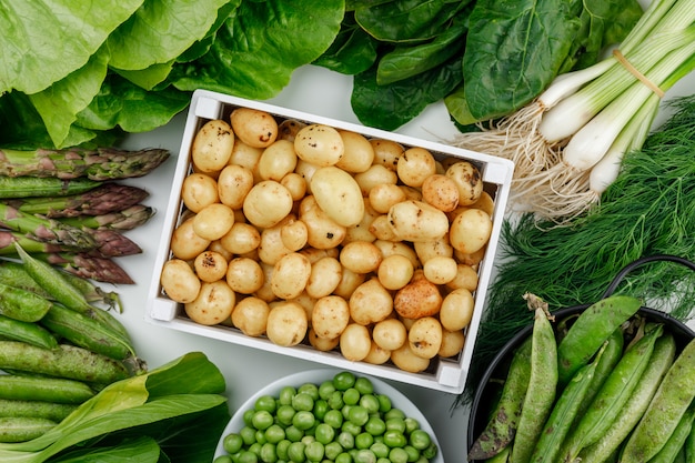 Patatas con vainas verdes, guisantes, eneldo, cebollas verdes, espinacas, acedera, lechuga, espárragos en una caja de madera en la pared blanca, vista superior.