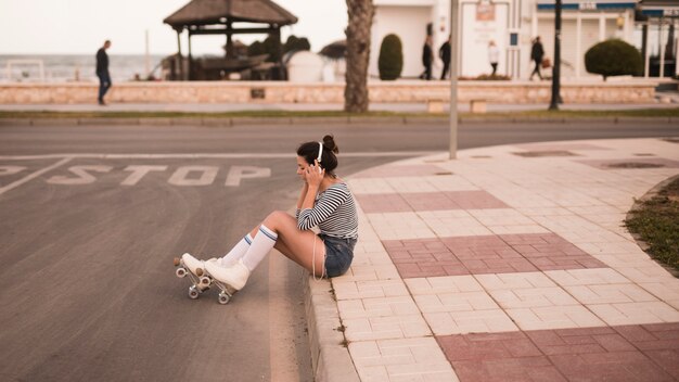 Patinador femenino joven que se sienta en música que escucha de la acera en el auricular