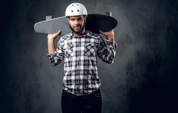 Un patinador negro vestido con una camisa de lana sostiene una tabla larga sobre un fondo gris en un estudio.