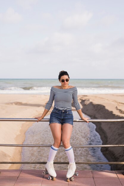 Patinador de sexo femenino joven de moda que se inclina en la verja cerca de la playa