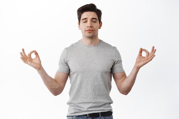 Paz dentro de ti. Hombre tranquilo y relajado respirando, meditando con los ojos cerrados y el rostro pacífico, manteniendo la mente bajo control, de pie contra el fondo blanco.