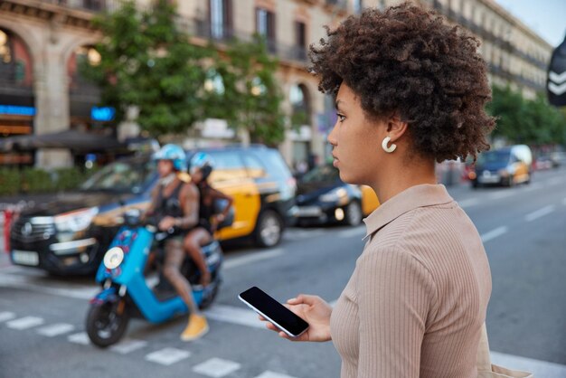 Peatonal femenino de pelo rizado cruza una carretera muy transitada tiene un teléfono inteligente moderno con poses de pantalla de maqueta en blanco al aire libre