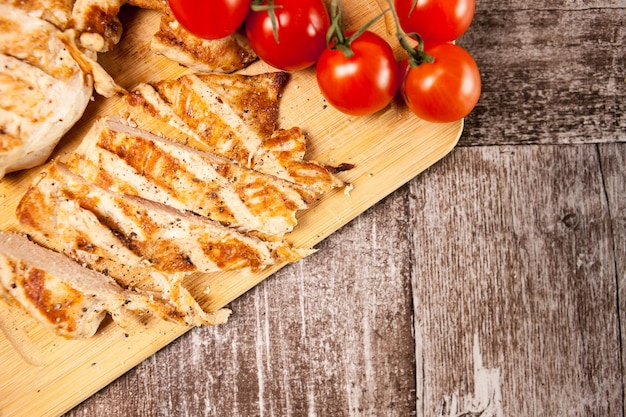 Pechuga de pollo fresca en rodajas a la parrilla sobre tabla de madera junto a tomates. Cena saludable y estilo de vida.