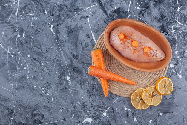 Pechuga de pollo en un recipiente junto a las zanahorias y el limón sobre un salvamanteles sobre la superficie azul