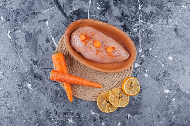 Pechuga de pollo en un recipiente junto a las zanahorias y el limón sobre un salvamanteles sobre la superficie azul