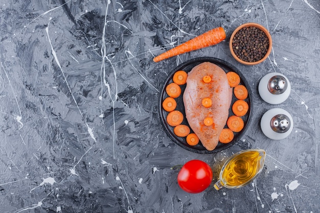 Pechuga de pollo y zanahorias en rodajas en un plato junto a sal, aceite, especias, zanahoria y tomate en la superficie azul