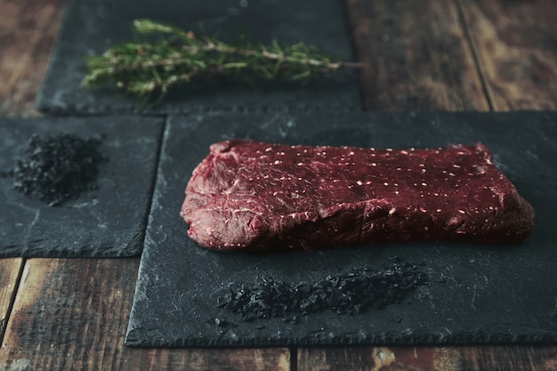 Pedazo de carne cruda en una almohadilla de piedra negra cerca de sal volcánica negra y hierbas y especias de romero