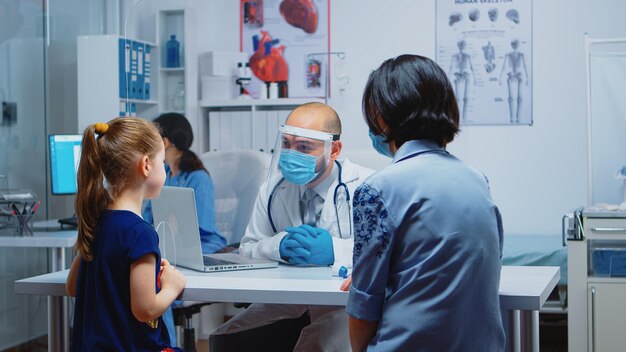 Pediatra explicando el tratamiento a la niña con máscara protectora. Especialista en medicina con mascarilla protectora brindando servicios de salud, consulta, tratamiento en hospital durante covid-19