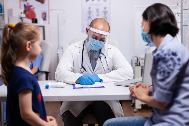 Pediatra interrogando al paciente sobre la enfermedad y escribiendo respuestas en el portapapeles. Médico en equipo de protección contra coronavirus discutiendo los síntomas en la clínica médica