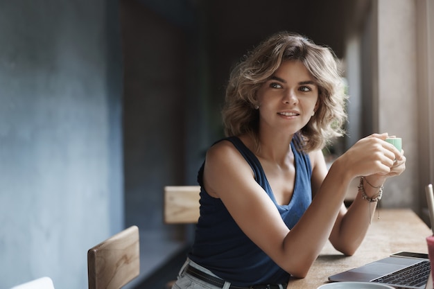 El peinado rizado rubio de la mujer joven moderna atrayente se aleja intrigado, sonriente, encantado, sostenga la taza, bebida, café, trabajando, computadora portátil, use net-book estudiando la tarea universitaria, investigando como autónomo.