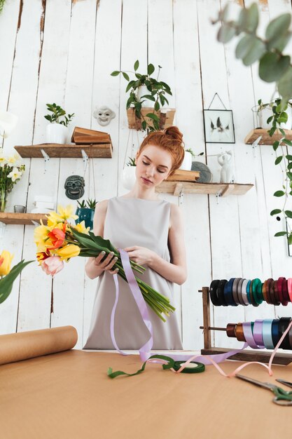 Pelirroja dama floristería recogiendo ramo mientras está de pie cerca de la mesa