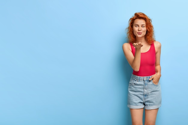 Pelirroja hermosa joven envía beso al aire, mira apasionadamente, expresa amor a sus seguidores, vestida con chaleco rojo y pantalones cortos de mezclilla, modelos contra la pared azul. Mwah y todo mi amor para ti.