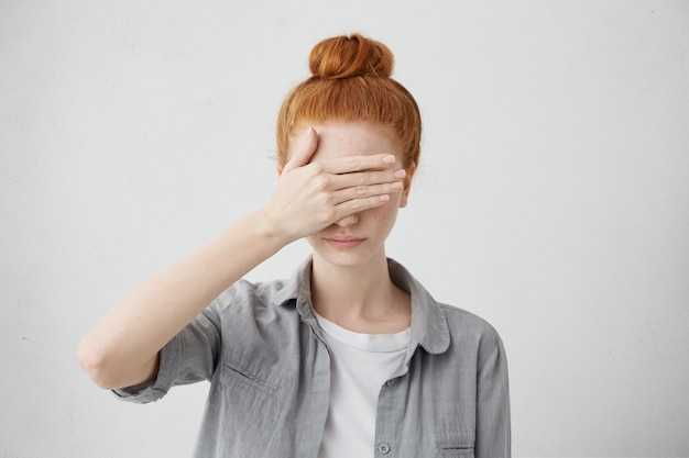 pelirroja con moño escondiendo los ojos debajo de la mano mientras se siente avergonzada. Mujer joven seria en ropa causal que cubre la cara con la mano. Expresiones y emociones faciales humanas.